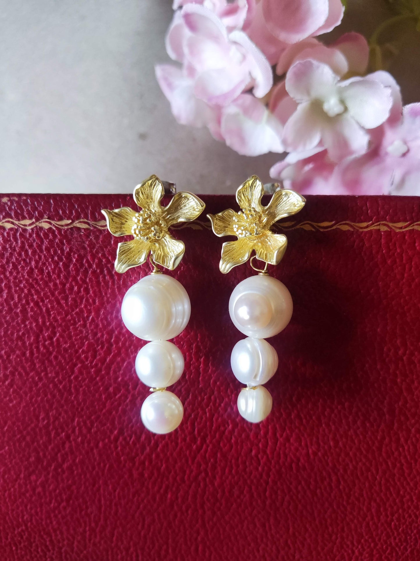 Pendientes dorados con flor y perlas de agua dulce sobre fondo rojo intenso.