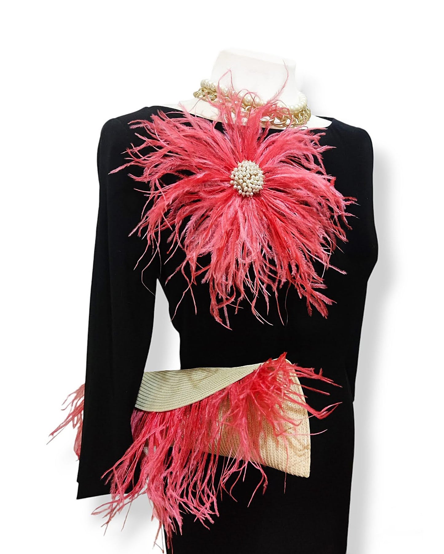 Maniquí con vestido negro llevando un clutch en tonos coral y beige, acompañado de un broche de plumas a juego.