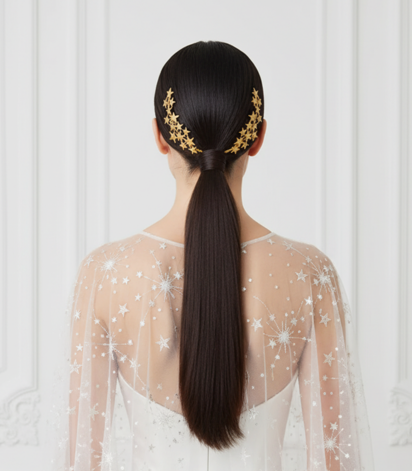 Bride wearing golden porcelain star hair combs on a sleek ponytail, photographed from the back against a white ornate wall.