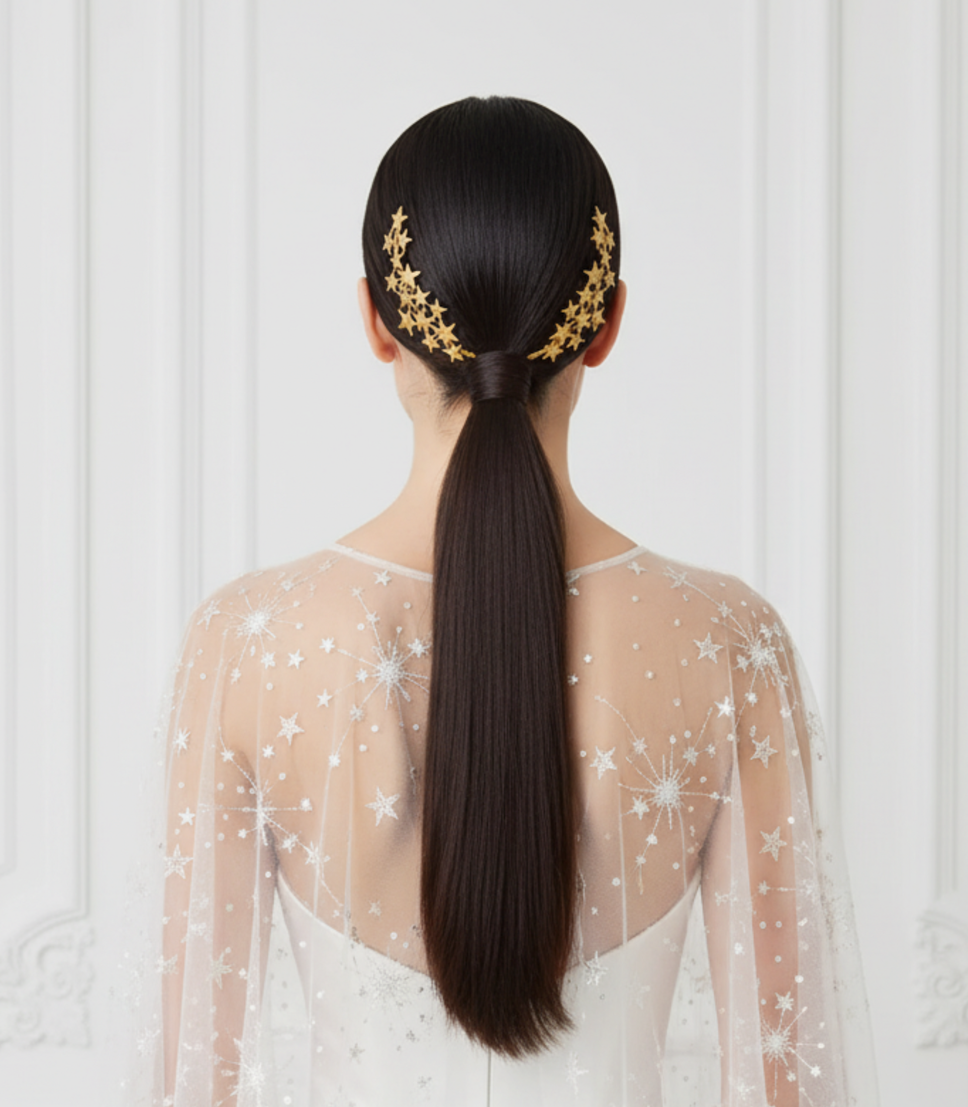 Bride wearing golden porcelain star hair combs on a sleek ponytail, photographed from the back against a white ornate wall.