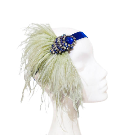 Diadema artesanal de terciopelo azul con aplique de cristal y plumas verde pistacho sobre fondo neutro.