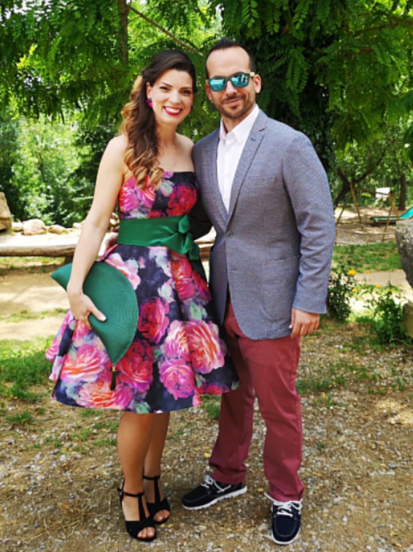 Pochette éventail vert bouteille tenue par une invitée de mariage portant une robe fleurie colorée. Accessoire élégant et artisanal signé Glo&Sons, parfait pour les cérémonies et événements estivaux.